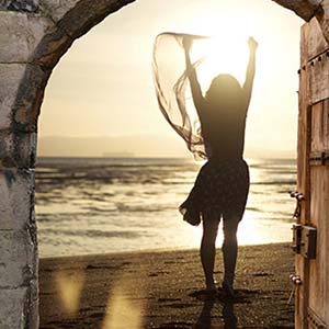 photo d'une porte ouverte donnant sur une plage où se trouve une personne les bras levés, face au soleil couchant